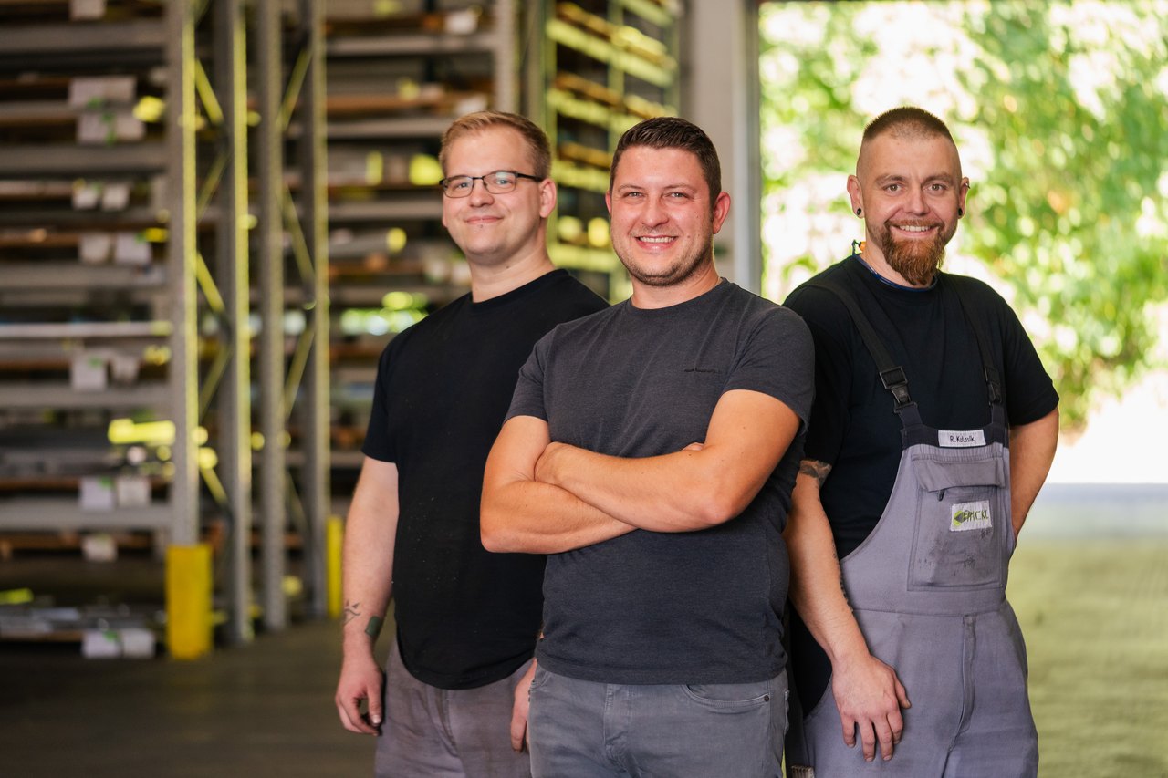 Drei lächelnde Männer in Arbeitskleidung stehen in einem Lagerhaus mit Metallregalen.