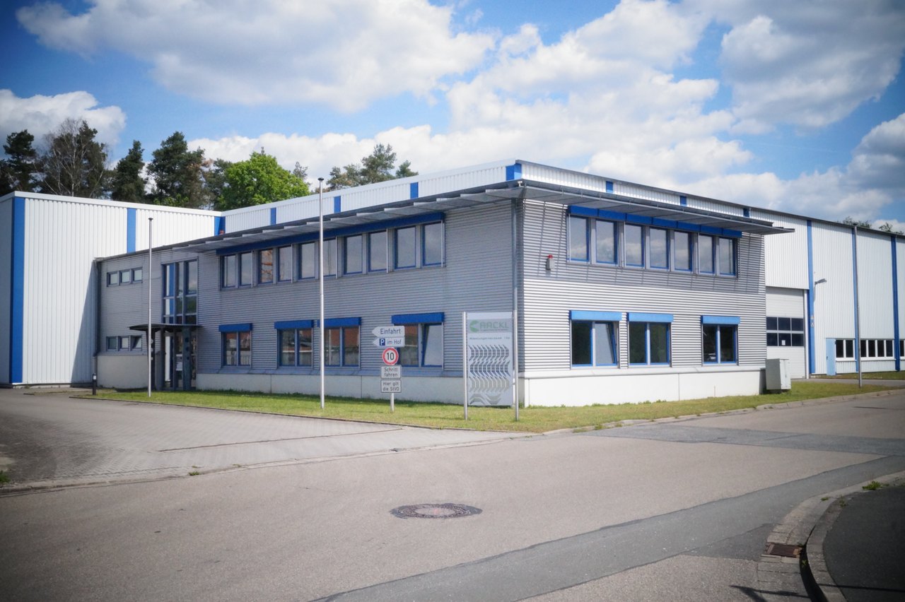 Ein graues, zweistöckiges Bürogebäude mit blauen Fensterrahmen und einem flachen Dach, umgeben von einem Parkplatz.