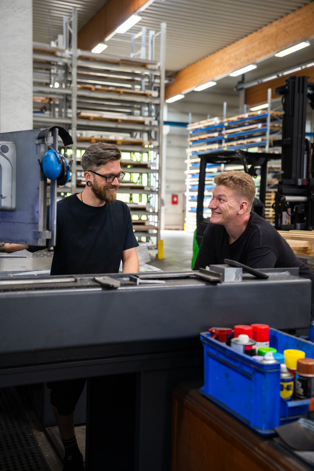 Zwei Männer lachen und unterhalten sich in einer Werkstatt, mit Regalen und Gabelstapler im Hintergrund.