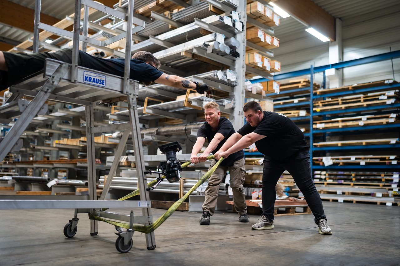 Zwei Arbeiter ziehen eine Plattform mit einem Mann darauf durch eine Lagerhalle voller Metallregale.