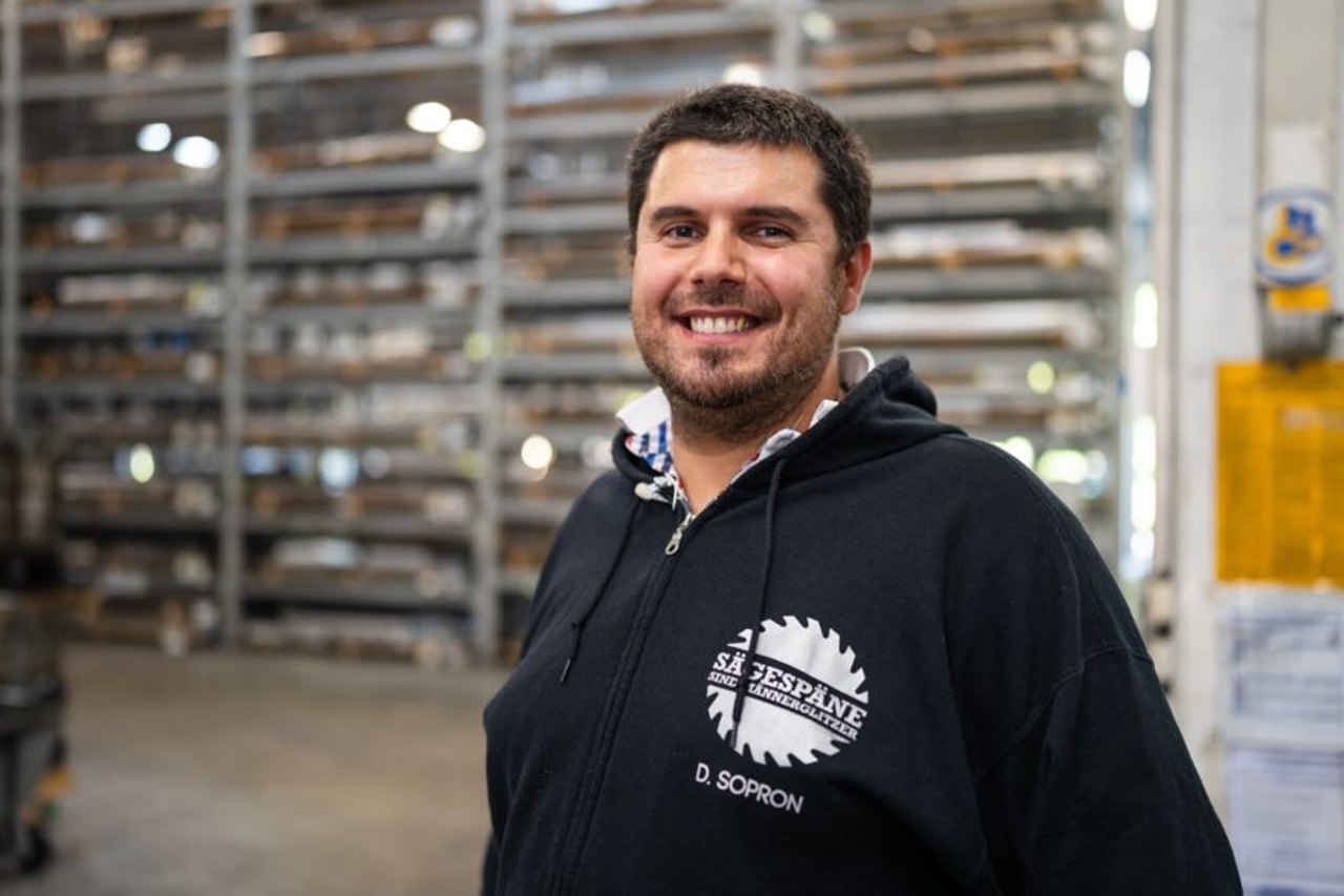 Lächelnder Mann in schwarzem Kapuzenpullover mit Logo "SZESZPAN D. SOPRON" vor Holzregalen.