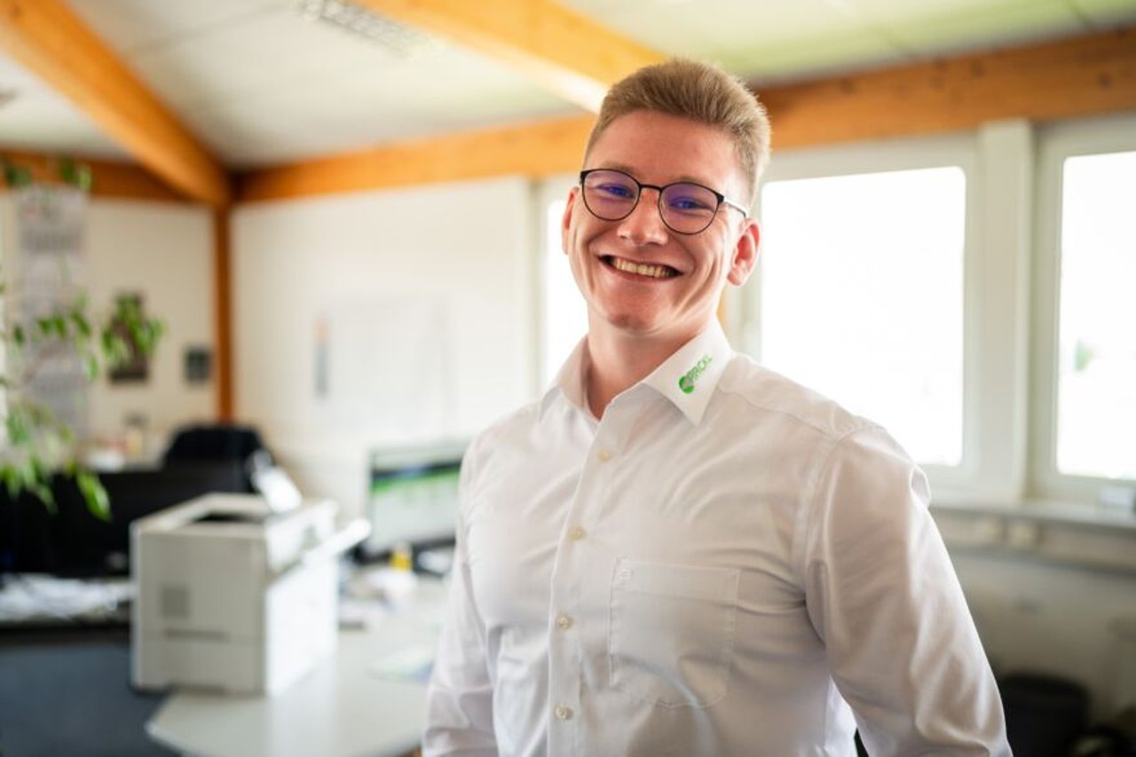 Lachender junger Mann mit Brille und weißem Hemd mit grünem Logo im Büro.