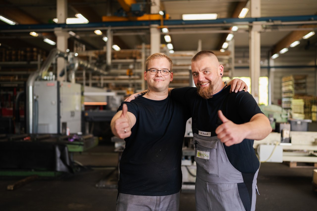 Zwei Männer in Arbeitskleidung, Arm in Arm, geben in einer Fabrikhalle den Daumen hoch.
