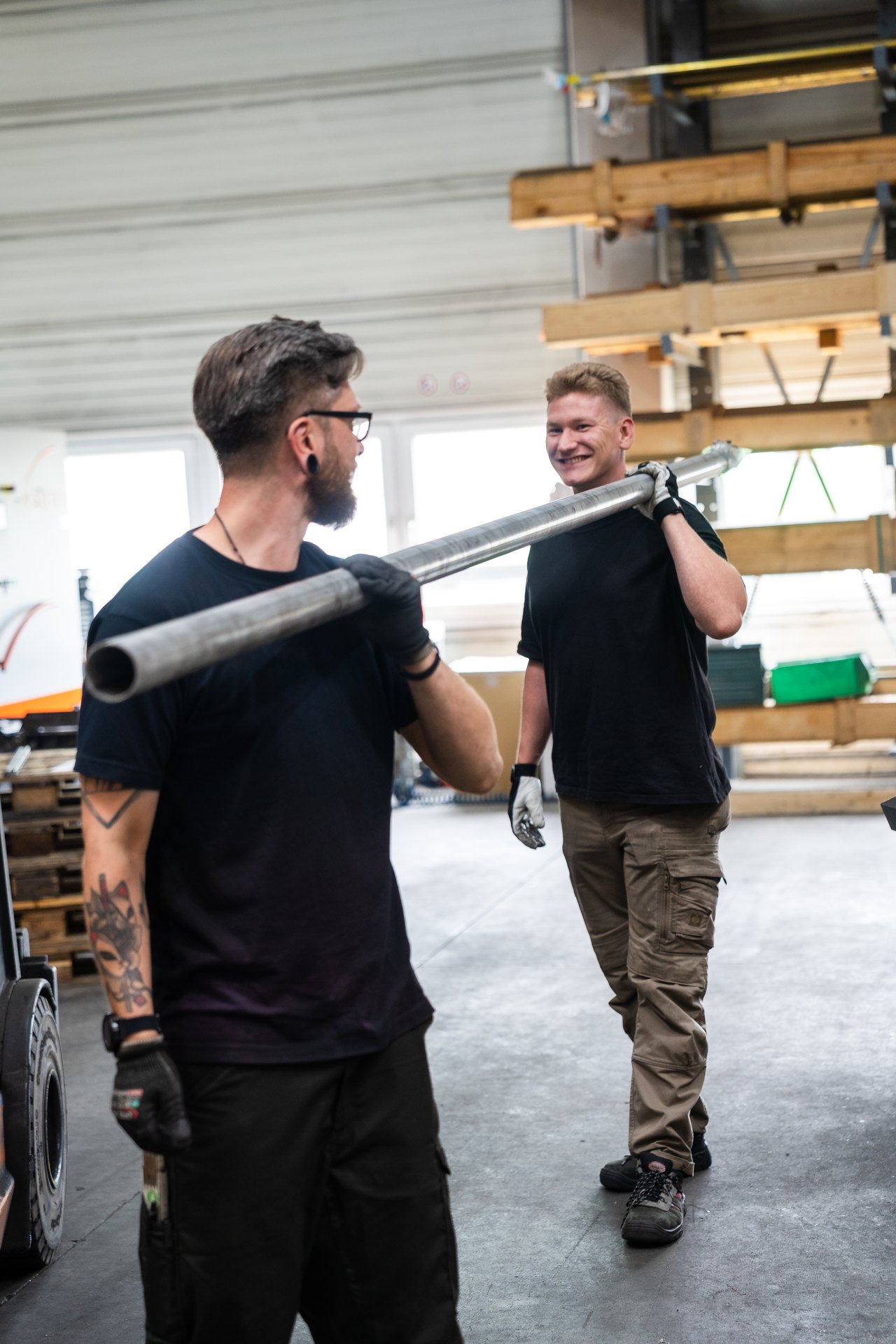 Zwei Männer in Arbeitshosen und T-Shirts tragen ein langes Metallrohr gemeinsam in einer Werkstatt.