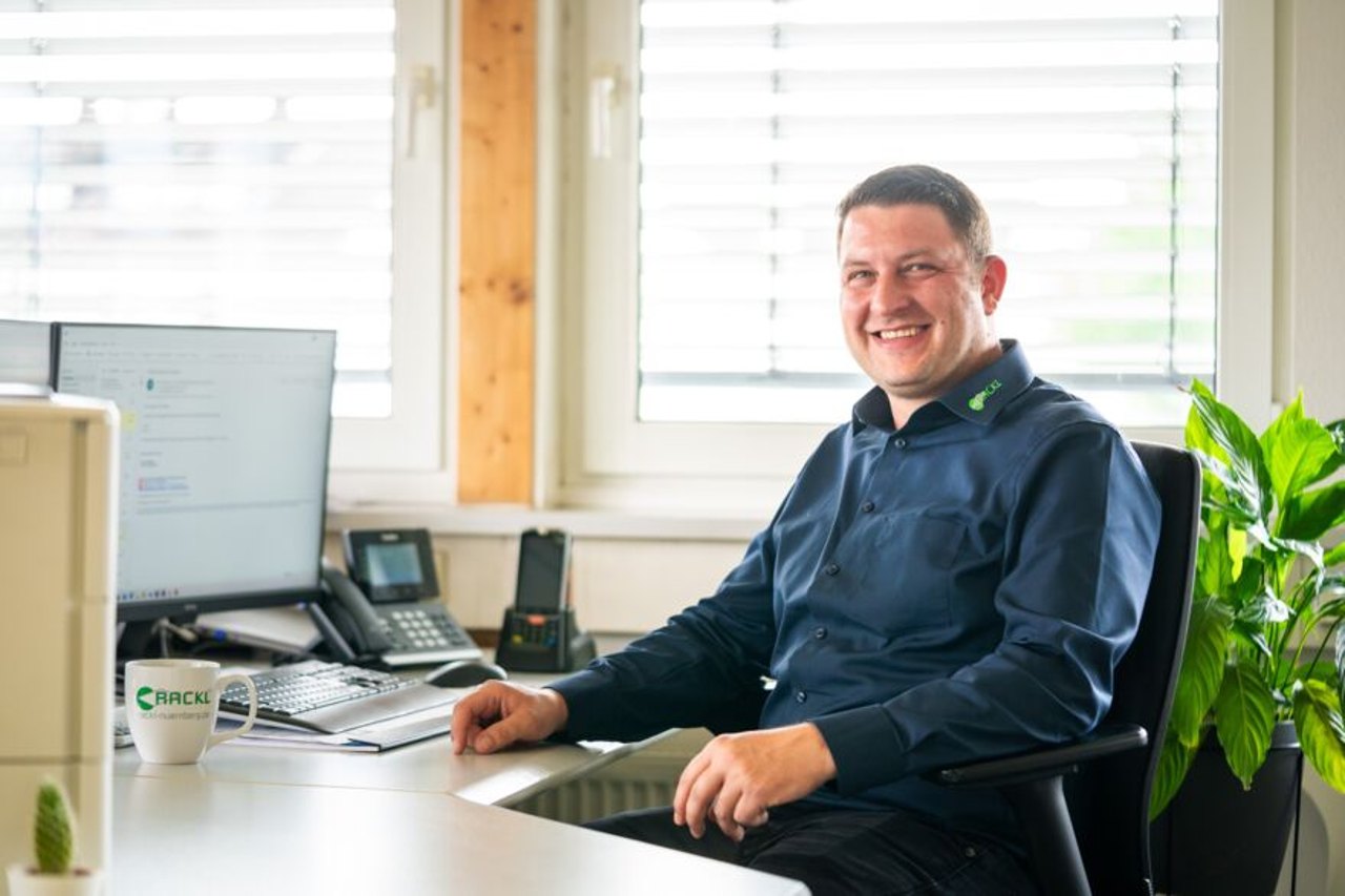 Ein lächelnder Mann in einem dunklen Hemd sitzt an einem Schreibtisch mit einem Computer und einer Tasse.