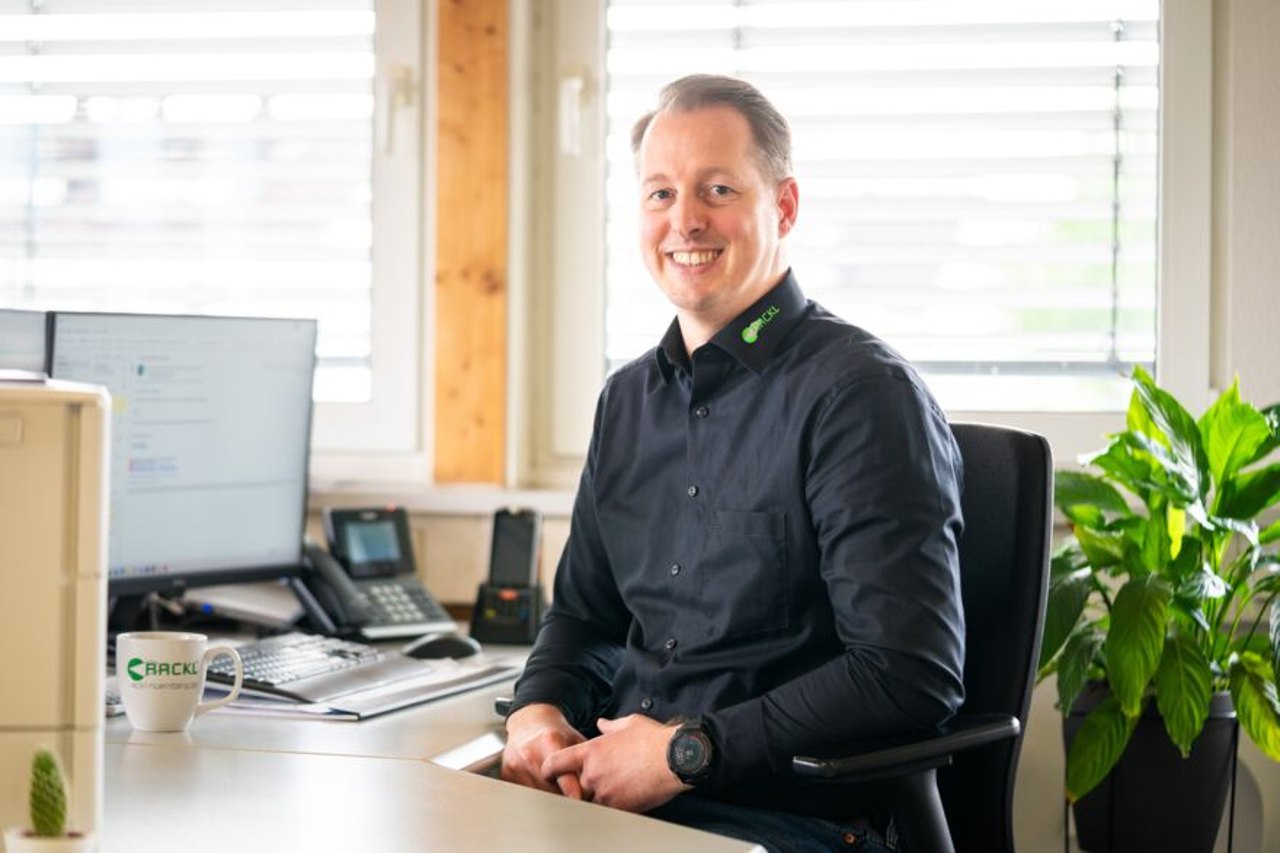 Ein lächelnder Mann sitzt an einem Bürotisch mit einem Computer und einer Tasse mit dem "EICKEL" Logo.