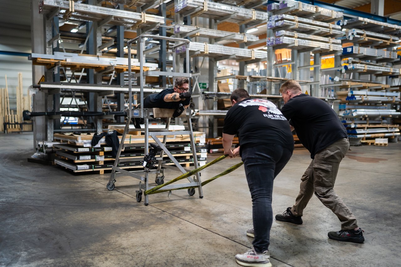 Zwei Männer ziehen ein Gerüst mit einem Mann darauf; Lagerregale im Hintergrund.