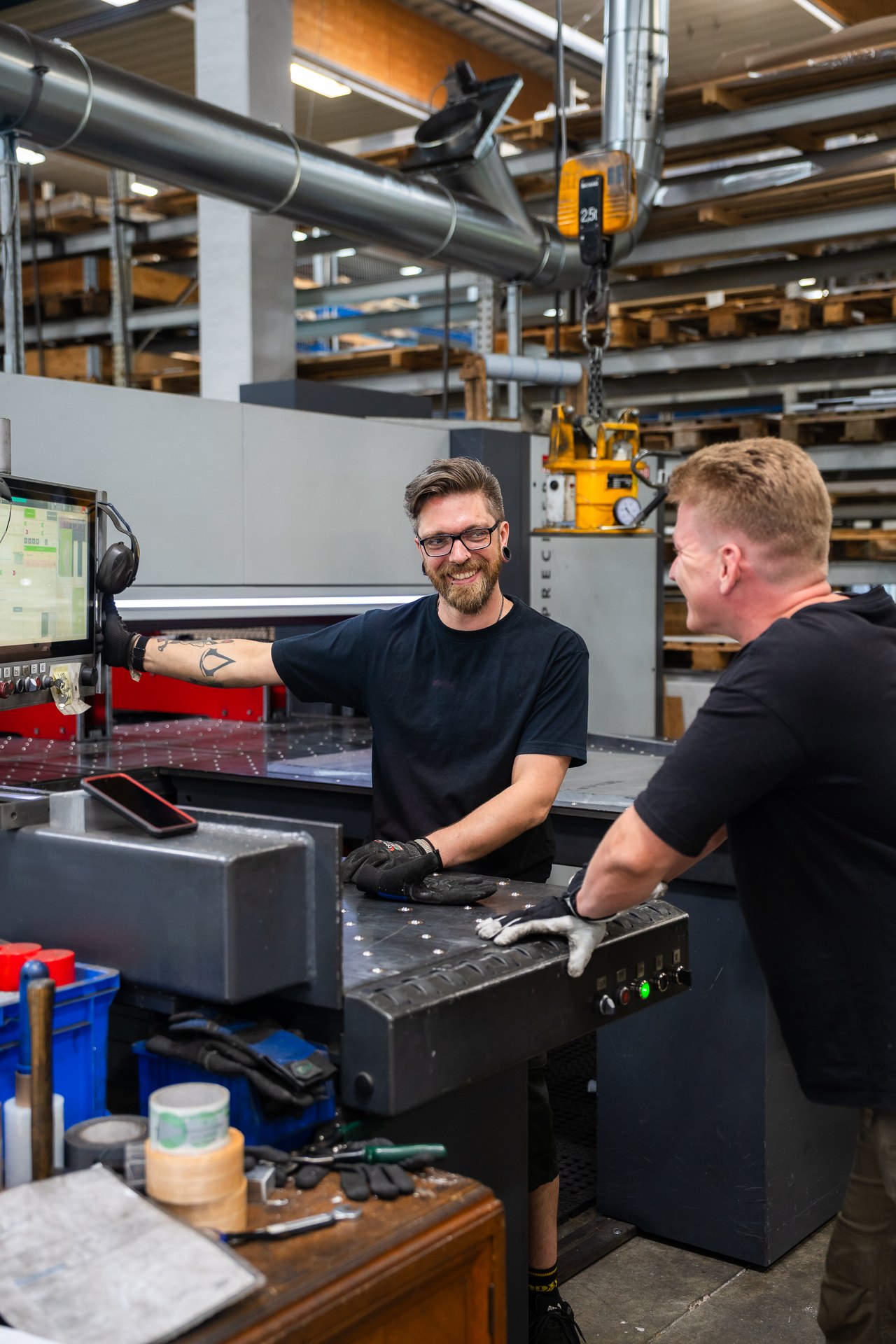 Zwei lächelnde Männer in schwarzen T-Shirts und Handschuhen arbeiten an Industriemaschinen.