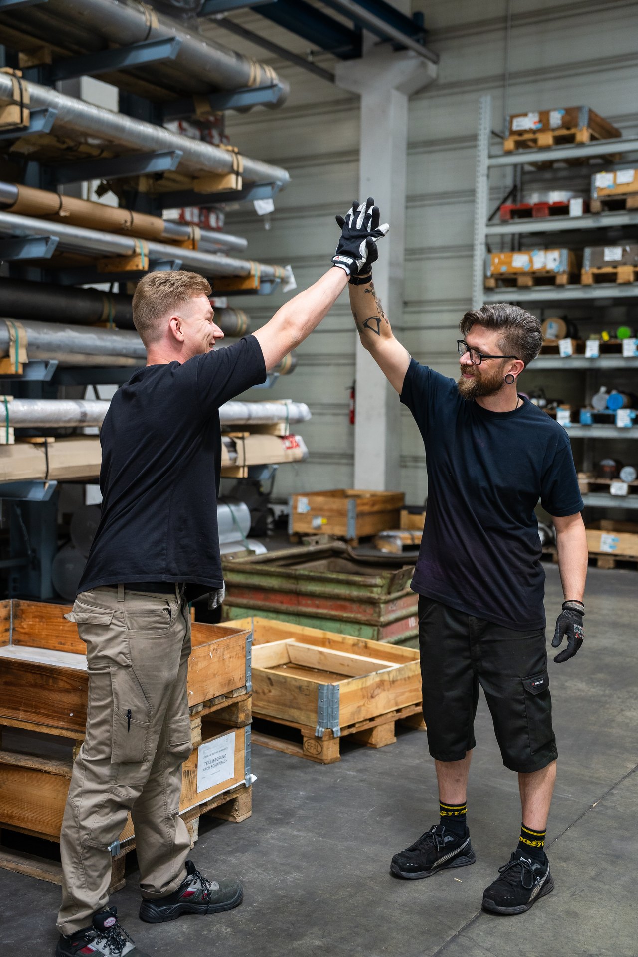Zwei lächelnde Männer geben sich im Lager ein High Five, umgeben von Regalen und Paletten.